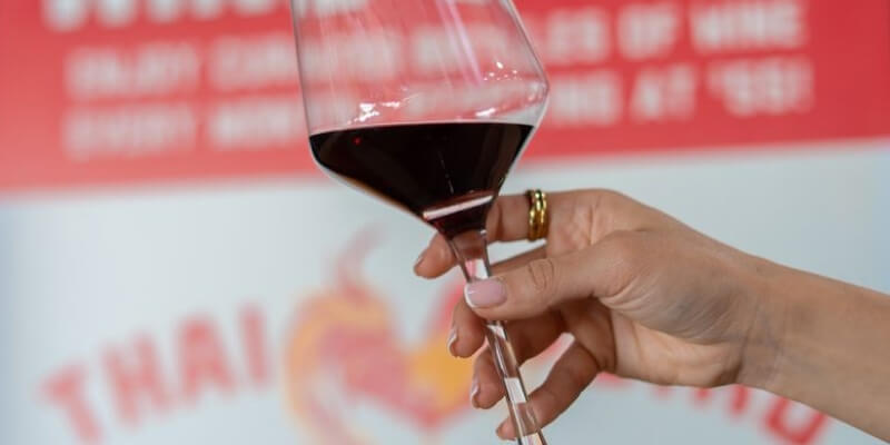 A person holding a glass of red wine in front of the Thai Bird Wine Club sign.