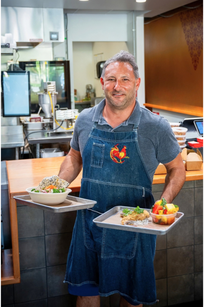 Smiling chef in a Thai Bird apron holding two trays of food in the restaurant.